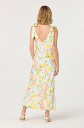 A woman is wearing a sleeveless, ankle-length dress with a watercolor floral pattern in pastel shades of yellow, pink, green, and blue. The dress features shoulder straps tied into bows on each shoulder and a deep V-shaped back. She is also wearing light-colored high-heeled shoes.